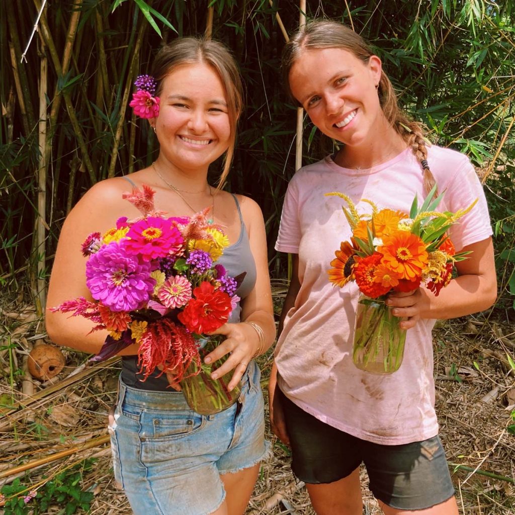 2 young ladies are smiling and standing in front of a bamboo forest. They are holding colorful flower bouquets. 