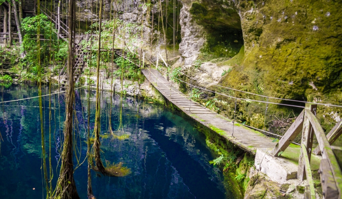 A wooden hanging bridge is on the side of the cliff to cross over the cenote's blue water.