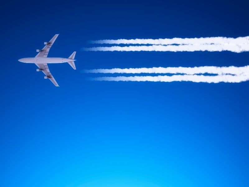 White plane is flying in a blue sky and leaving white lines pictured from below. 