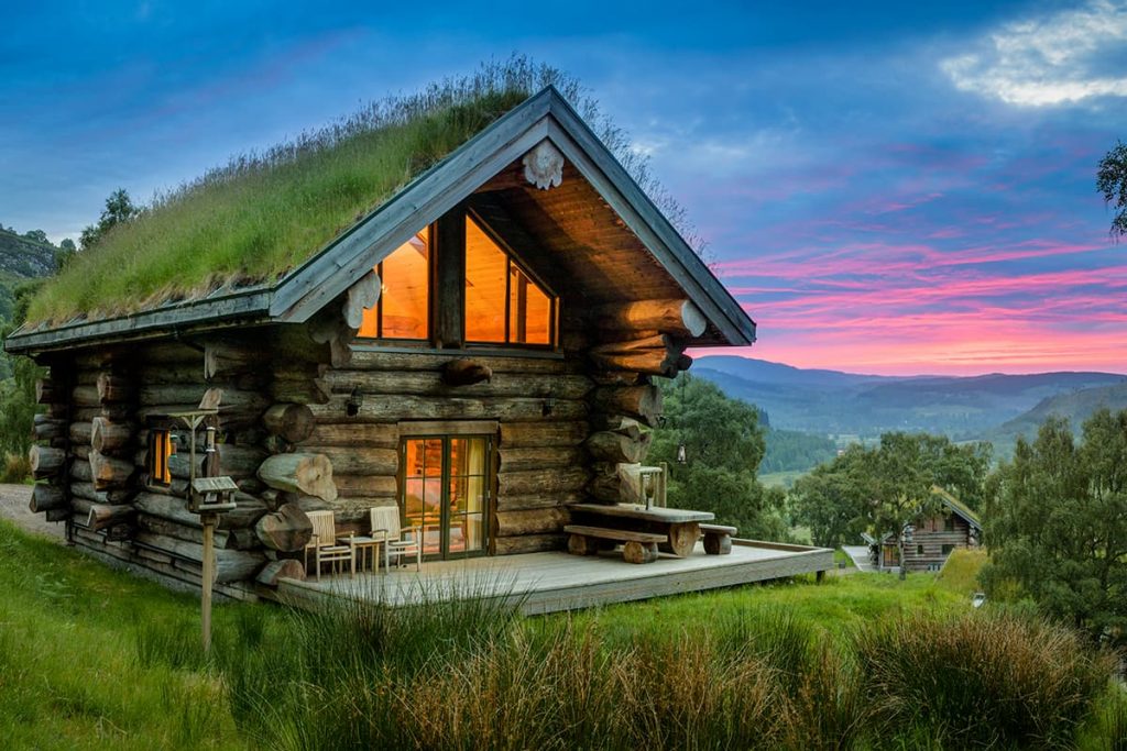 Small wooden log cabin with grass roof on top of a hill with a sunset behind it