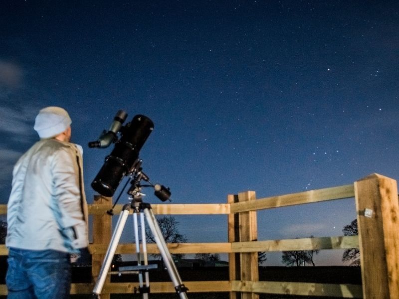 Woman with a telescope is watching the night sky behind a fence
