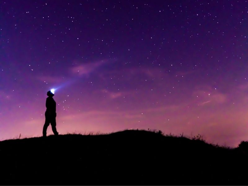 A dark figure with a flashlight in front of a purple pink sky with stars