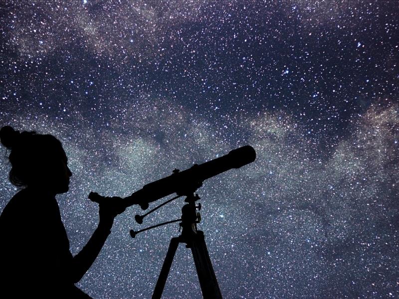 A dark figure with a telescope with starry night above