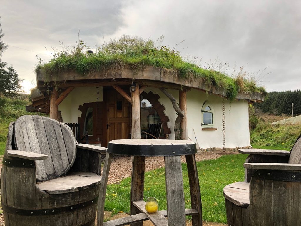 Small cottage with grass roof and white walls and wooden stilts. Reclaimed wood chairs in the foreground