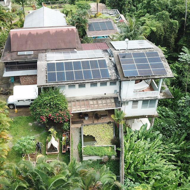 A white grey building from above with lush green vegetarian, solar panels on the roof and a garden with small pond. 