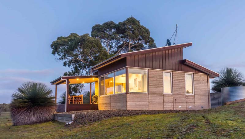 A small wooden hut with a front porch situated on top of a hill. The lights are turned on. 