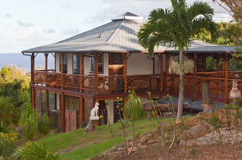 2 storey house with 360 degree wooden porch on a hillside with grey roof. 