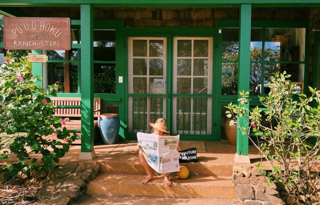 Green wooden house with a porch and a girl is sitting on the steps and reading a newspaper. Wooden sign says Puu O Hoku Ranch Store