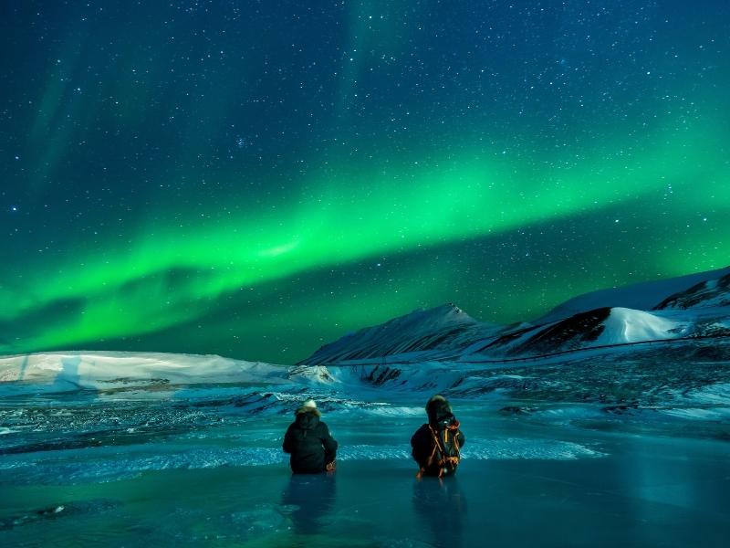 2 people with backpacks and thick clothing is standing in icy water and watching the green Northern lights