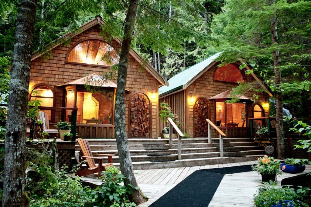 2 cut wooden cottages right next to each other in the middle of a pine forest with lights on. 