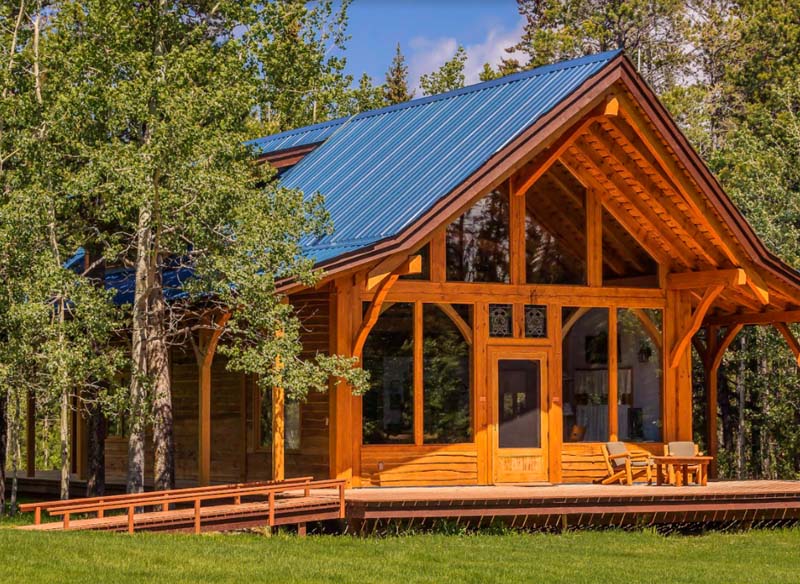 A wooden lodge with large windows to the front, a small porch and silver roof surrounded by trees. 