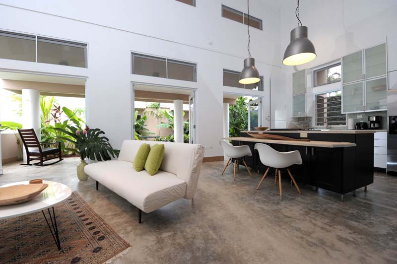 Interior of a house with white walls and large windows. There are light brown furnitures and dark brown kitchen island. 