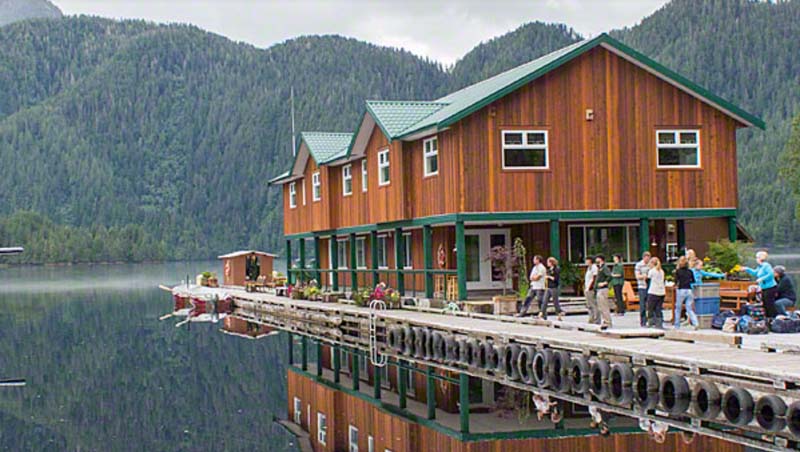 A brown and green wooden two-storey house that is floating on a pier in the middle of the lake. People are walking along it. 