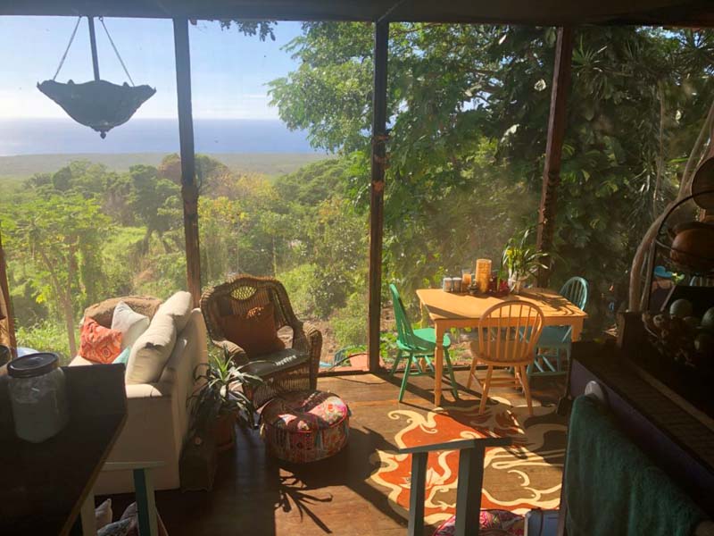 A room with a ceiling to floor large windows overlooking a green forest. There are several furnitures and colorful decoration. 