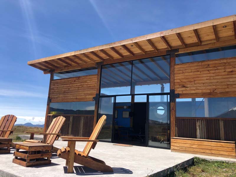 A small wooden cottage with huge glass windows and doors with a concrete terrace in the front with wooden chairs. 