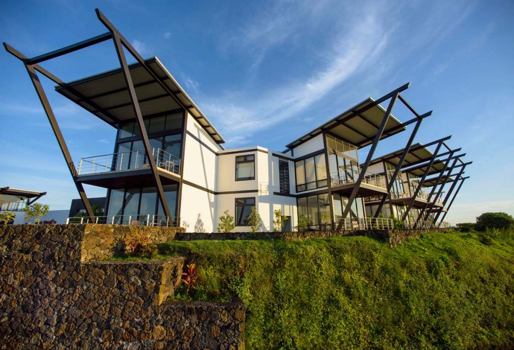 White lodge with huge glass windows and black wooden shades on top of a green hill