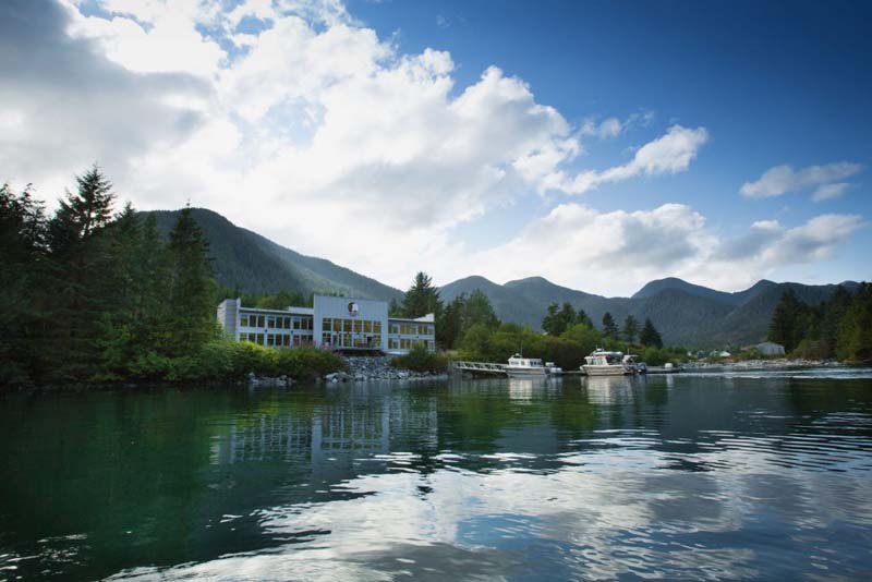 A silver two-storey house right at the riverbank with boats anchored in front of it. Surrounded by huge mountains. 