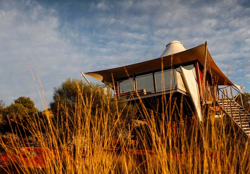 A small house on stilts above a red sandy ground. Stairs lead up to the side and the house is covered with canvas instead of classic roof. 