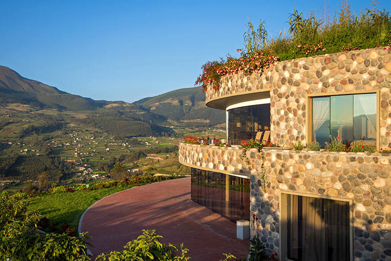 A round-shaped two-storey house covered with round stones and a huge red terrace in the front overlooking a valley with small houses. 