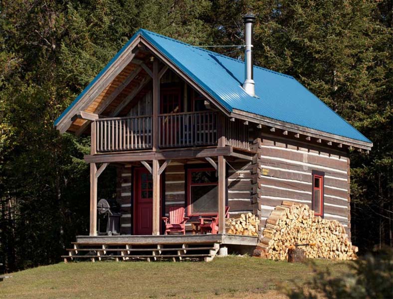 Small wooden house with blue roof, red windows and door and a small front porch. Chopped up firewood is stacked on one side. 