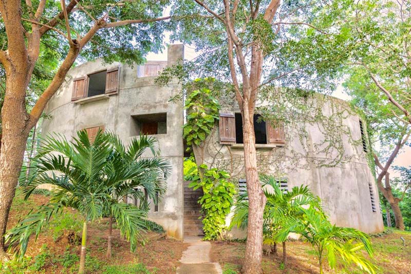 Round shaped grey house made of concrete surrounded by green trees and plants