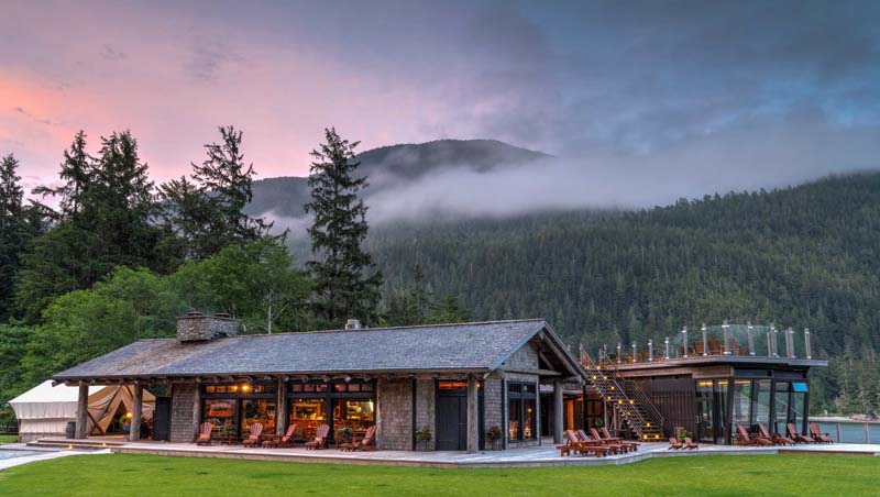 A wooden lodge right next to a lake with a large forest and mountain in the background. 