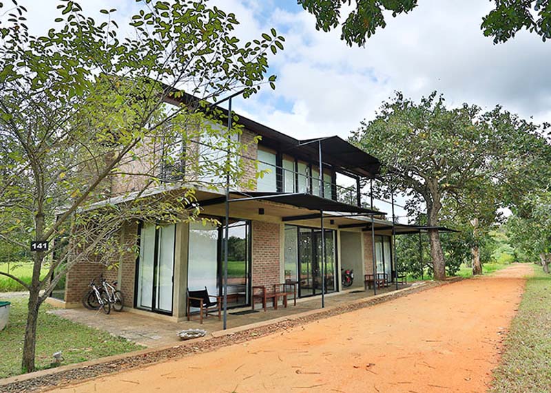 Two storey building made of bricks and metal roof with large windowes and doors next to a sandy road. 