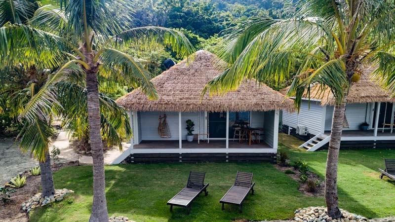 A white wooden hut with a roof made of palm leaves and a front porch in the middle of a rainforest. 