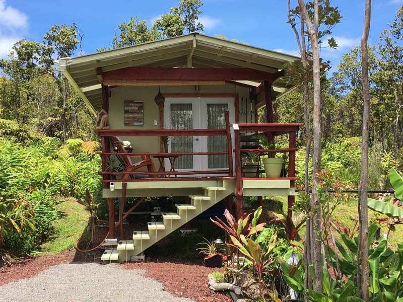 Small green and brown hut on stilts with a small front porch and large white doors surrounded by green plants. 