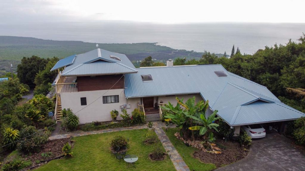 A house on the hillside overlooking the ocean surrounded by grass, pavements and several green plants and trees. 