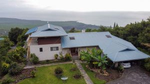 Large white house with light blue roof overlooking the ocean and the mountain side.