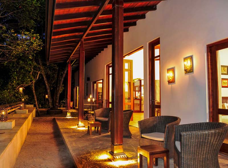 White one storey building with wooden roof and large doors as windows. There is a patio in the front with woven chairs and small tables