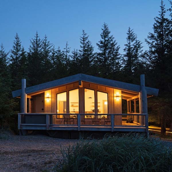 Small wooden cottage with large windows and circular porch surrounded by trees. 