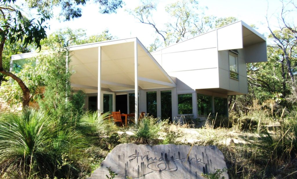 A modern white building with lots of windows and doors and a front terrace covered with a white roof. 