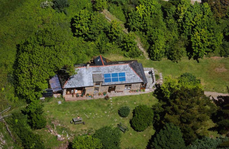 Aerial view of a two story brick house with solar panels on the roof laying in the middle of a green field and forest. 