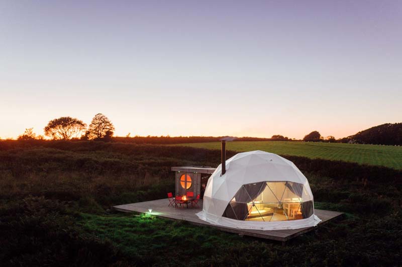 Small white dome-like tent one side of which is opaque. It is erected in the middle of a green field where nothing is around it except for grass. It has a small wooden hut and a terasse. 