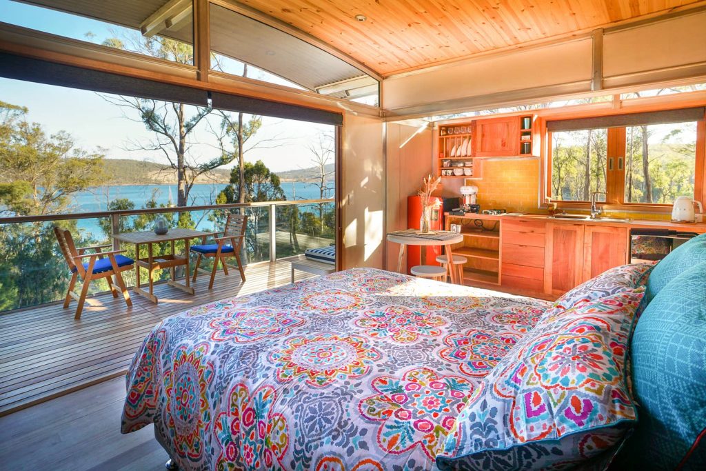 Double bed with colorful duvet and a small wooden kitchen next to it. The huge glass door is left open to the terrace so you can see the ocean view. 