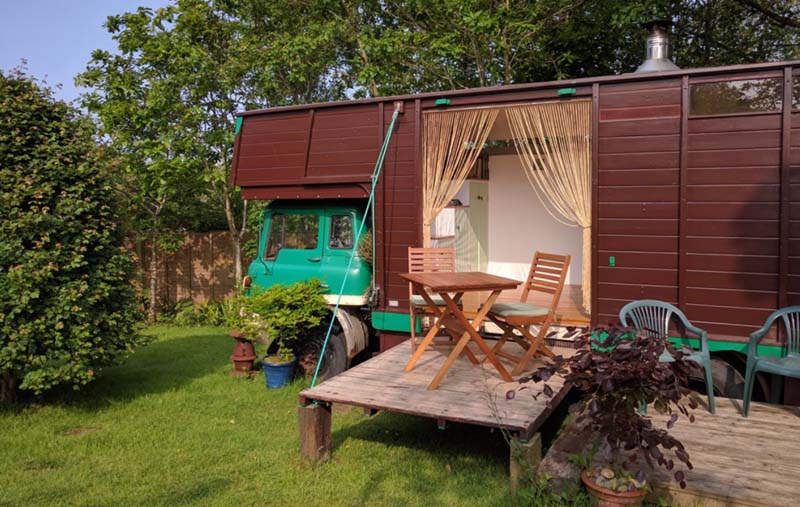 A green old truck with a dark brown wooden construction at the back which has a large door to go it. It has a small terrace with 2 chairs and a small table in the front. 