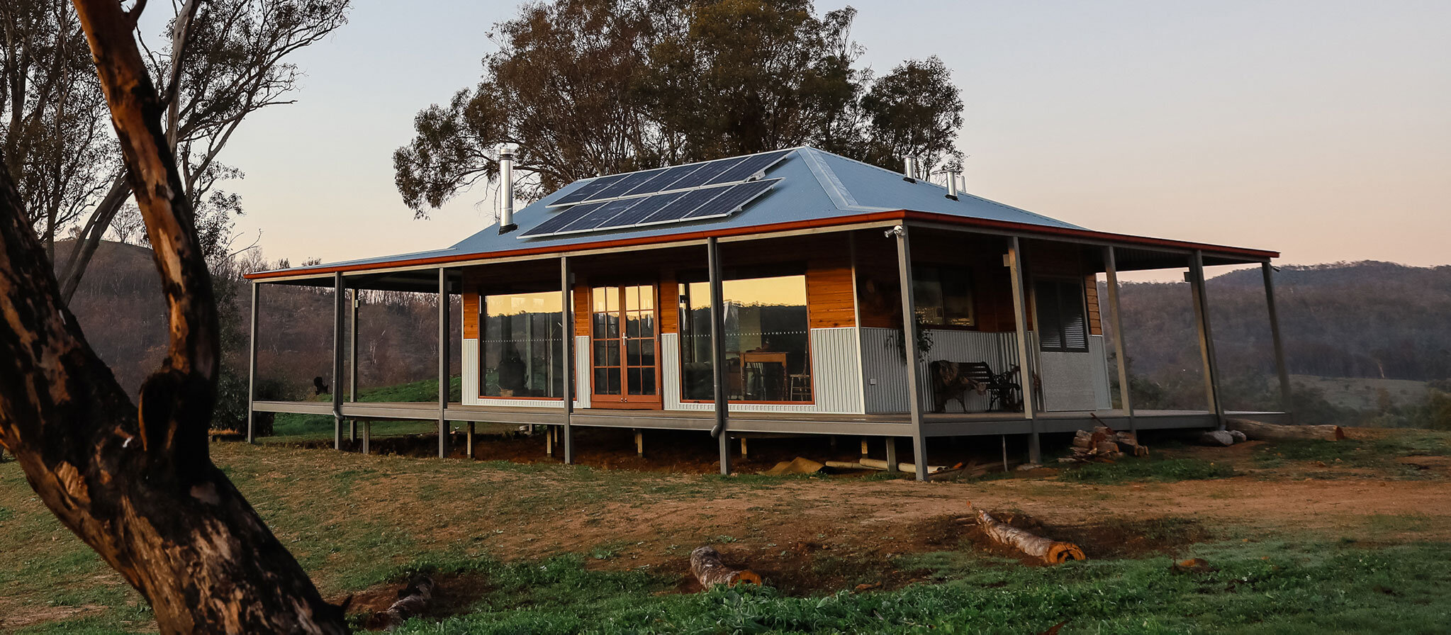 One-storey small wooden hut with solar panels on the metal roof and a porch around the whole hut. 