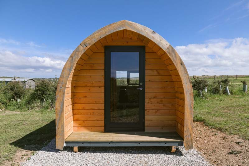 Small wooden dome-shaped pod in gravel with a door facing towards us. It is in the middle of a green field with nothing in sight