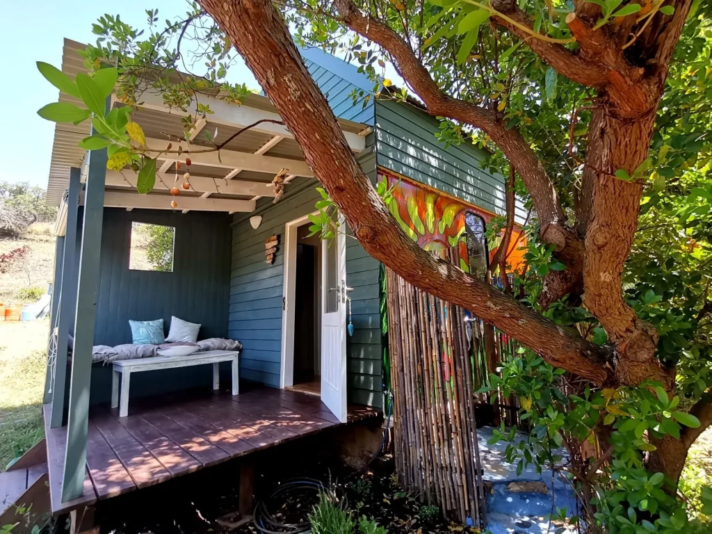 It is a small wooden cottage painted green and white with a small outdoor deck. It is surrounded by trees. 