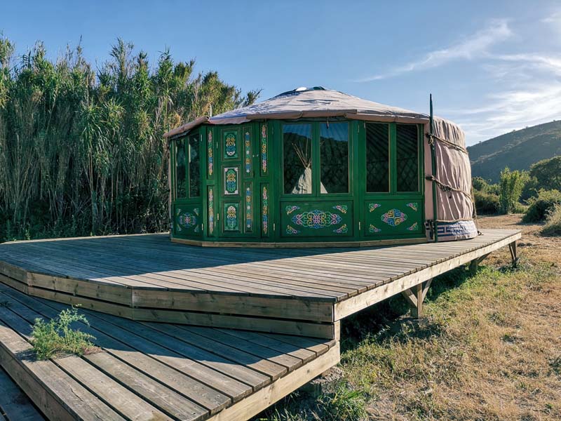 A wooden deck with a round-shaped yurt. Its windows and door and painted green with colorful floral patterns. 