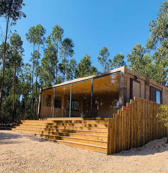 A small wooden cottage with sliding doors covering one side completely. Stairs are leading up to the doors. It is surrounded by trees. 