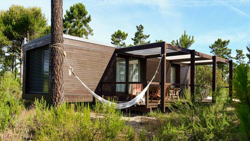 Small dark wooden cabin with a small outdoor deck surrounded by trees. Large windows are on the side and it is standing on small stilts without a permanent base. 