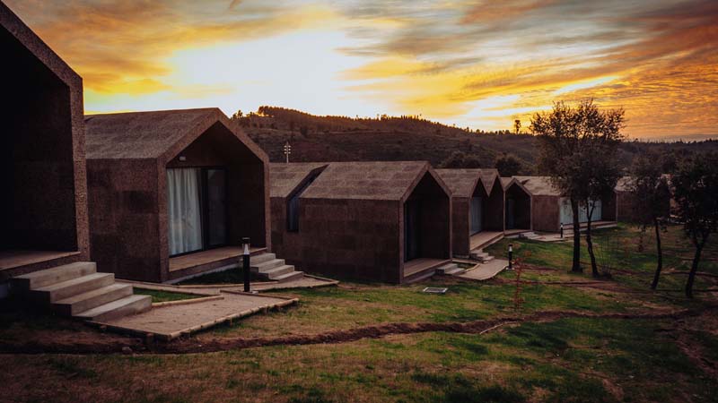 Several small buildings made of cork right next to each other on a hill side. A sun is setting behind them. 