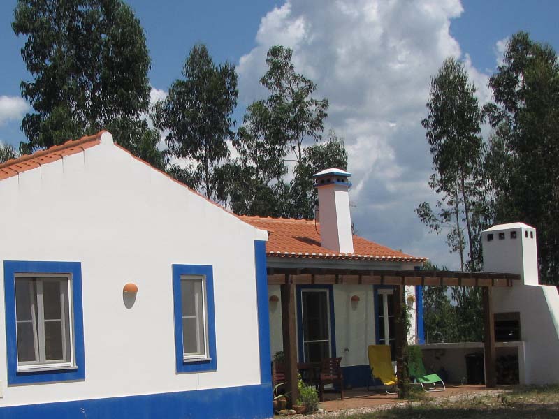 White and vibrant blue house with a terracotta roof. It has a patio with chair and an outdoor oven. 