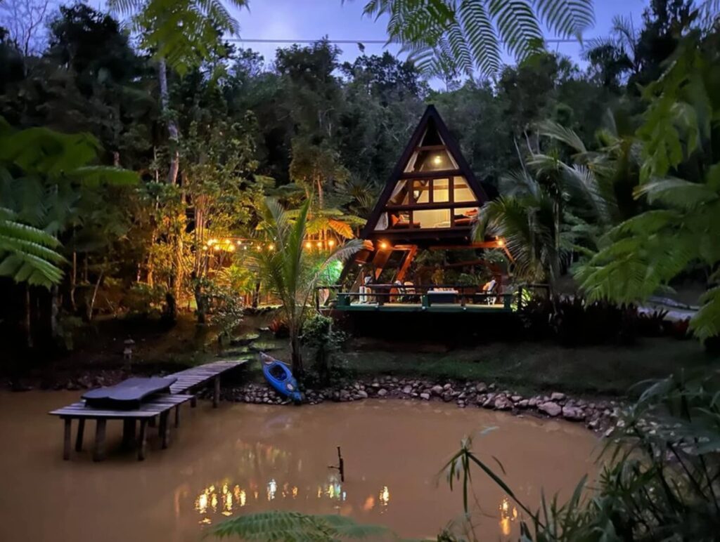 A-shaped hut in the jungle with a lake in front of it. 