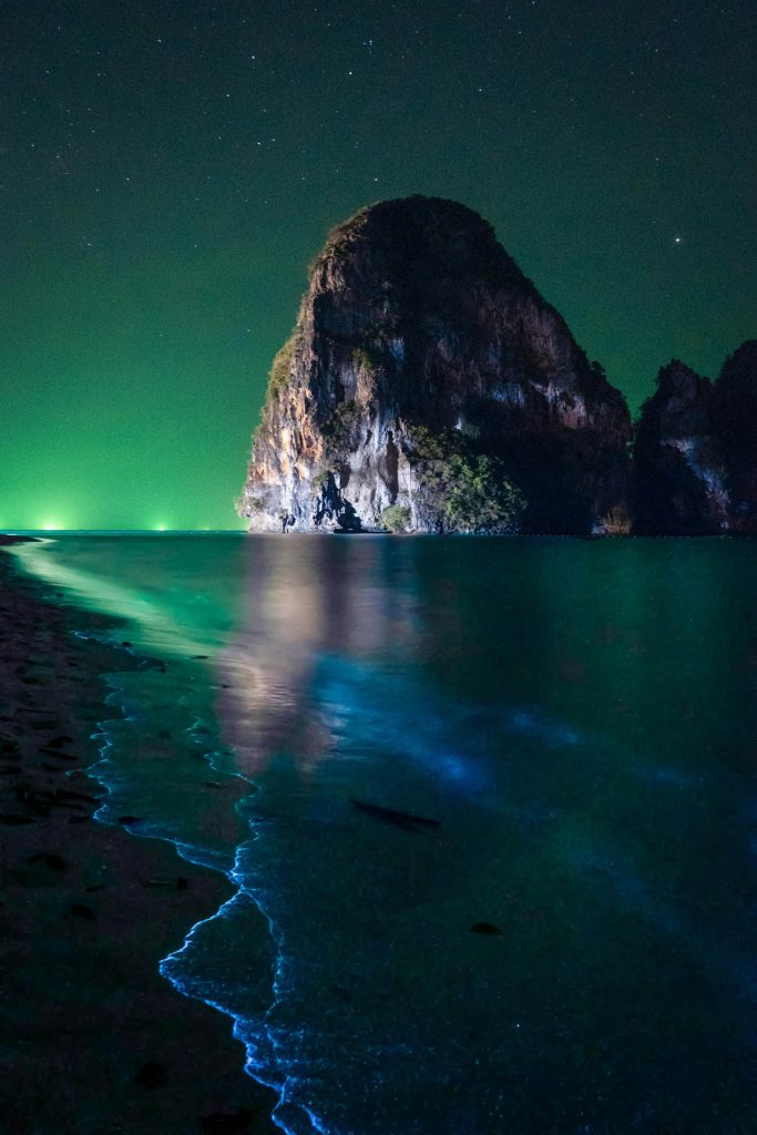 Sea at night with fluorescent blue and green swirls. Rocky hill is at the background.