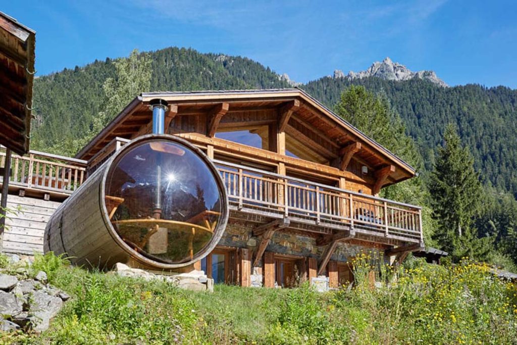 A two-storey wooden house with a long balcony. A small round shaped glass and wood structure is in front f=of it.