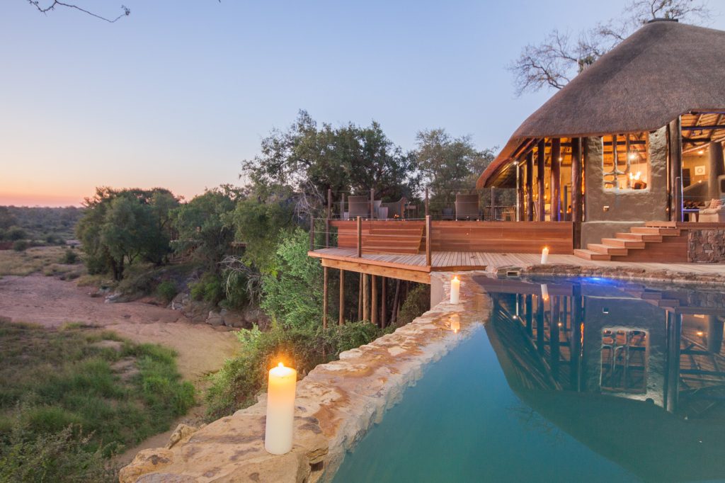 A wooden house with straw bale roof, huge balcony and a pool right in front of it.